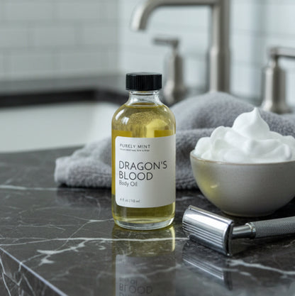 A bottle of Dragon's Blood body oil by Purely Mint on a dark marble bathroom counter next to a small bowl of shaving cream, a metal razor, and a gray towel, with a blurred modern bathroom sink in the background.