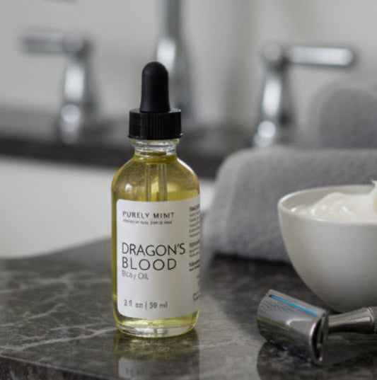 A bottle of Dragon's Blood body oil by Purely Mint on a dark marble bathroom counter next to a small bowl of shaving cream, a metal razor, and a gray towel, with a blurred modern bathroom sink in the background.