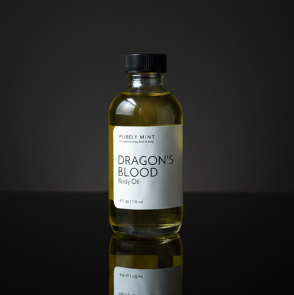 A bottle of Dragon's Blood body oil by Purely Mint, featuring a clear glass bottle filled with golden-yellow oil, a white label, and a black cap, set against a dark reflective background.