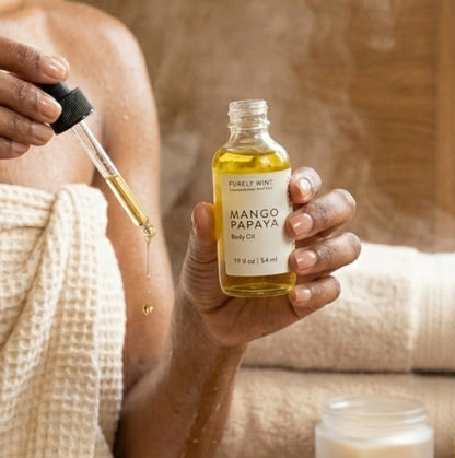 A woman wrapped in a beige waffle towel holds a small glass bottle of Mango Papaya body oil with a dropper, with steam rising from behind the bottle. A folded towel, a small jar, and a sponge sit on a wooden table in a spa-like setting.