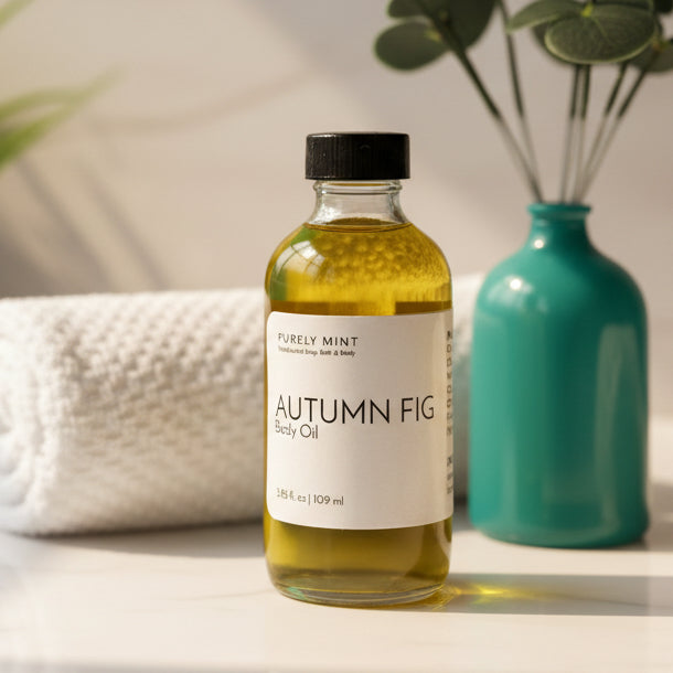 Bottle of 'Autumn Fig' body oil with a white label on a light surface, next to a teal vase and green plant and white rolled washcloth.