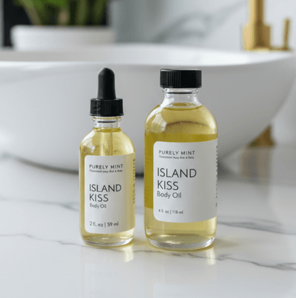 Two glass bottles of Purely Mint Island Kiss Body Oil standing on a white marble surface with a blurred white sink and gold faucet in the background. One bottle is a smaller dropper bottle and the other is a larger bottle with a black cap.