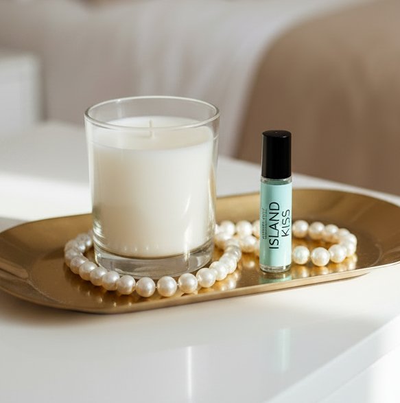 Island Kiss perfume oil sits on a gold tray with a pearl bracelet next to a white candle in a glass jar, set on a white bedside table in a softly lit bedroom.