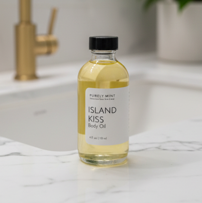 A bottle of Purely Mint Island Kiss Body Oil stands on a white marble countertop in front of a blurred bathroom sink and gold faucet. The clear bottle contains pale yellow oil and has a black cap with a white label.