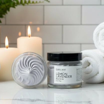 Lemon Lavender body butter in a luxury bathroom with rolled white washcloths, candles and a green plant on a marble counter. 
