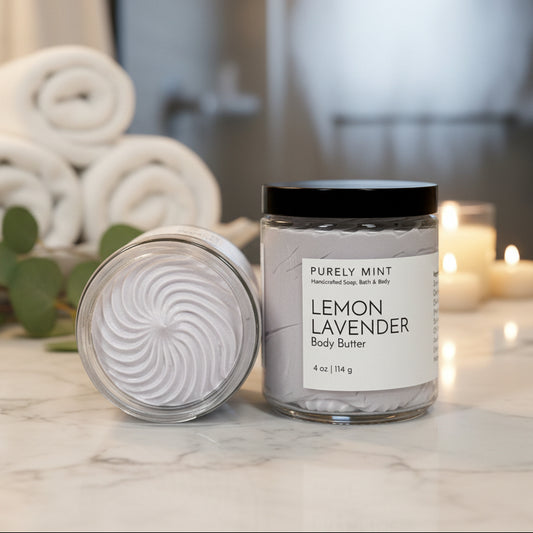 Lemon Lavender body butter in a luxury bathroom with rolled white washcloths, candles and a green plant on a marble counter. 