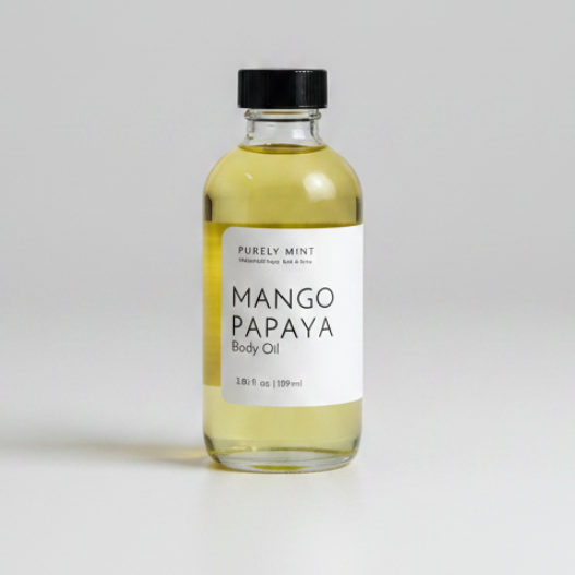 A bottle of Purely Mint Mango Papaya Body Oil with pale yellow oil inside, a white label, and a black cap, set against a neutral light gray background.