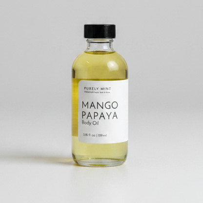 A bottle of Purely Mint Mango Papaya Body Oil with pale yellow oil inside, a white label, and a black cap, set against a neutral light gray background.