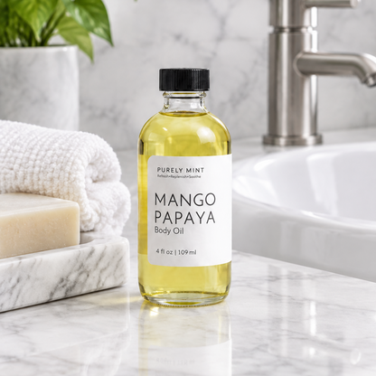 A 4 oz bottle of Purely Mint Mango Papaya Body Oil stands on a white marble bathroom counter next to a marble soap dish with a folded white towel and bar of soap, with a modern bathroom sink in the background.