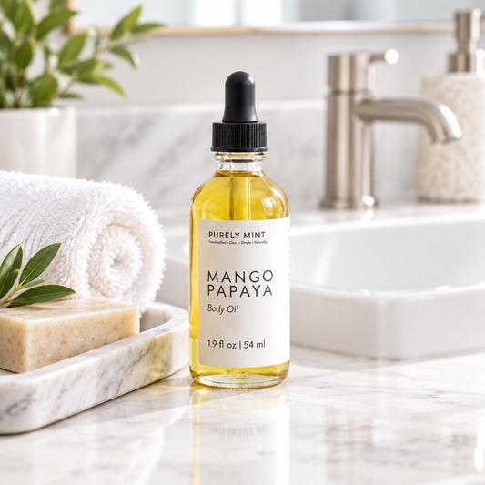 A 2 oz bottle of Purely Mint Mango Papaya Body Oil stands on a white marble bathroom counter next to a marble soap dish with a folded white towel and bar of soap, with a modern bathroom sink in the background.
