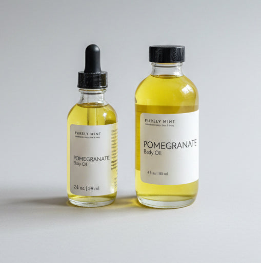 Two small glass bottles of Pomegranate Body Oil with white labels and black caps, one with a dropper and one with a cap, sitting on a neutral gray backdrop.
