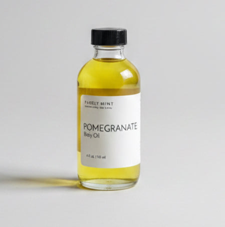 A small bottle of Pomegranate Body Oil with a black cap and white label sits on a neutral gray backdrop.