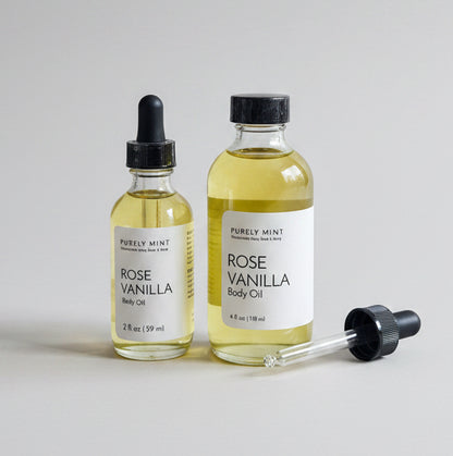 Two glass bottles of Rose Vanilla Body Oil with black dropper caps, standing on a light gray seamless backdrop.