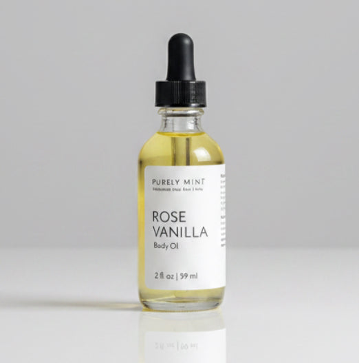 A 2 oz glass dropper bottle of Rose Vanilla Body Oil with a white label and black dropper cap, placed on a light gray backdrop. 