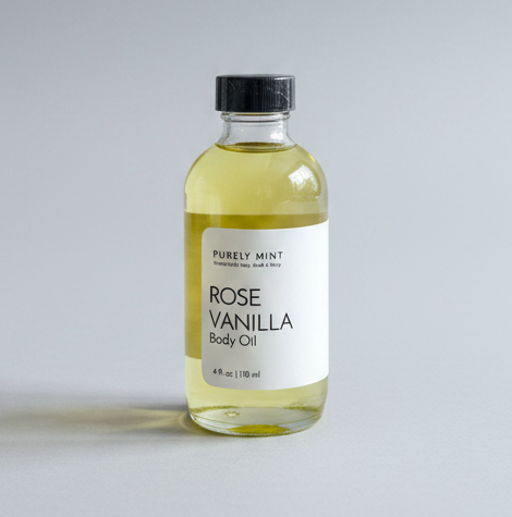 A 2 oz glass dropper bottle of Rose Vanilla Body Oil with a white label and black dropper cap, placed on a light gray backdrop. 