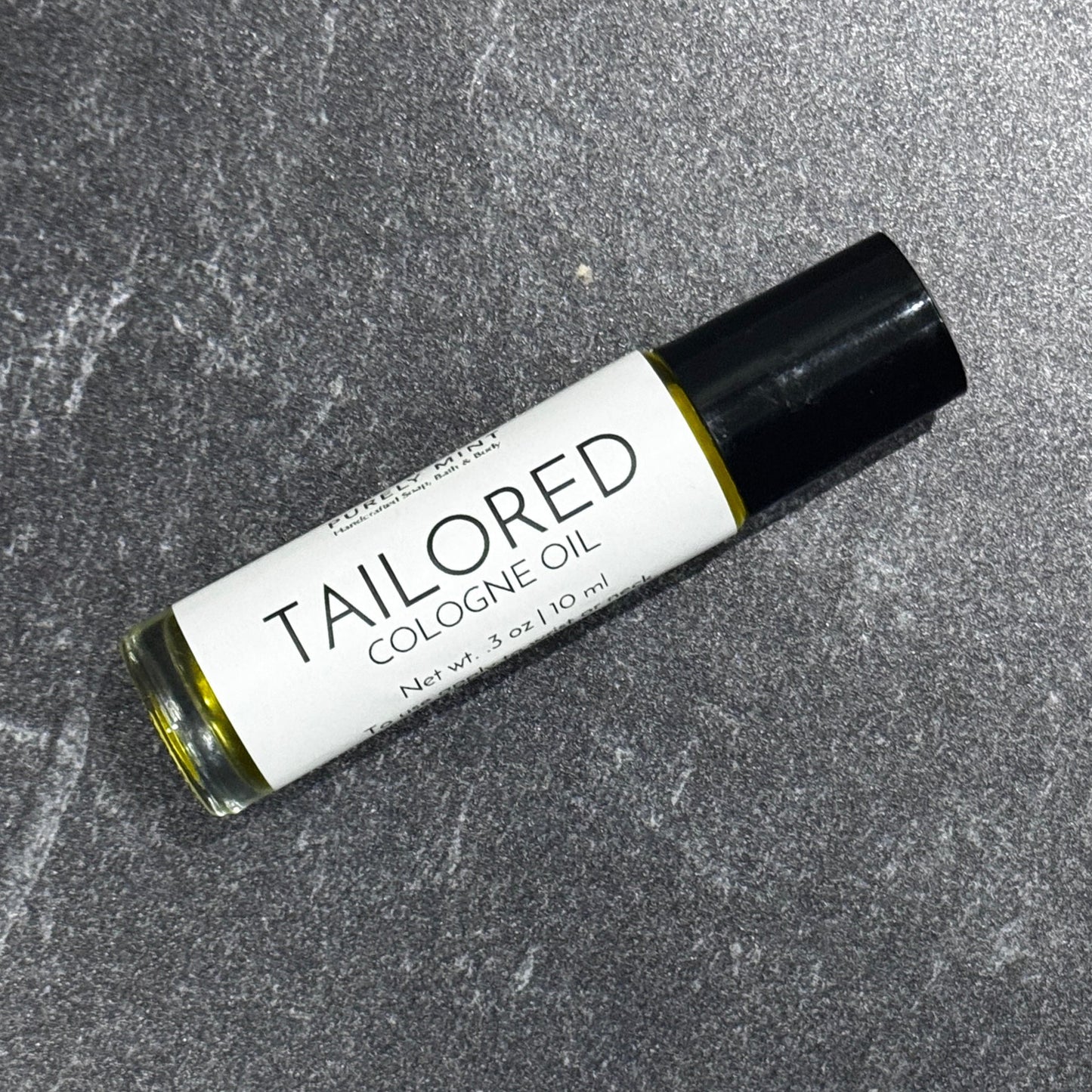 A 10 ml roll-on bottle of Tailored Cologne Oil by Purely Mint, featuring a white label and black cap, resting on a textured gray stone surface.