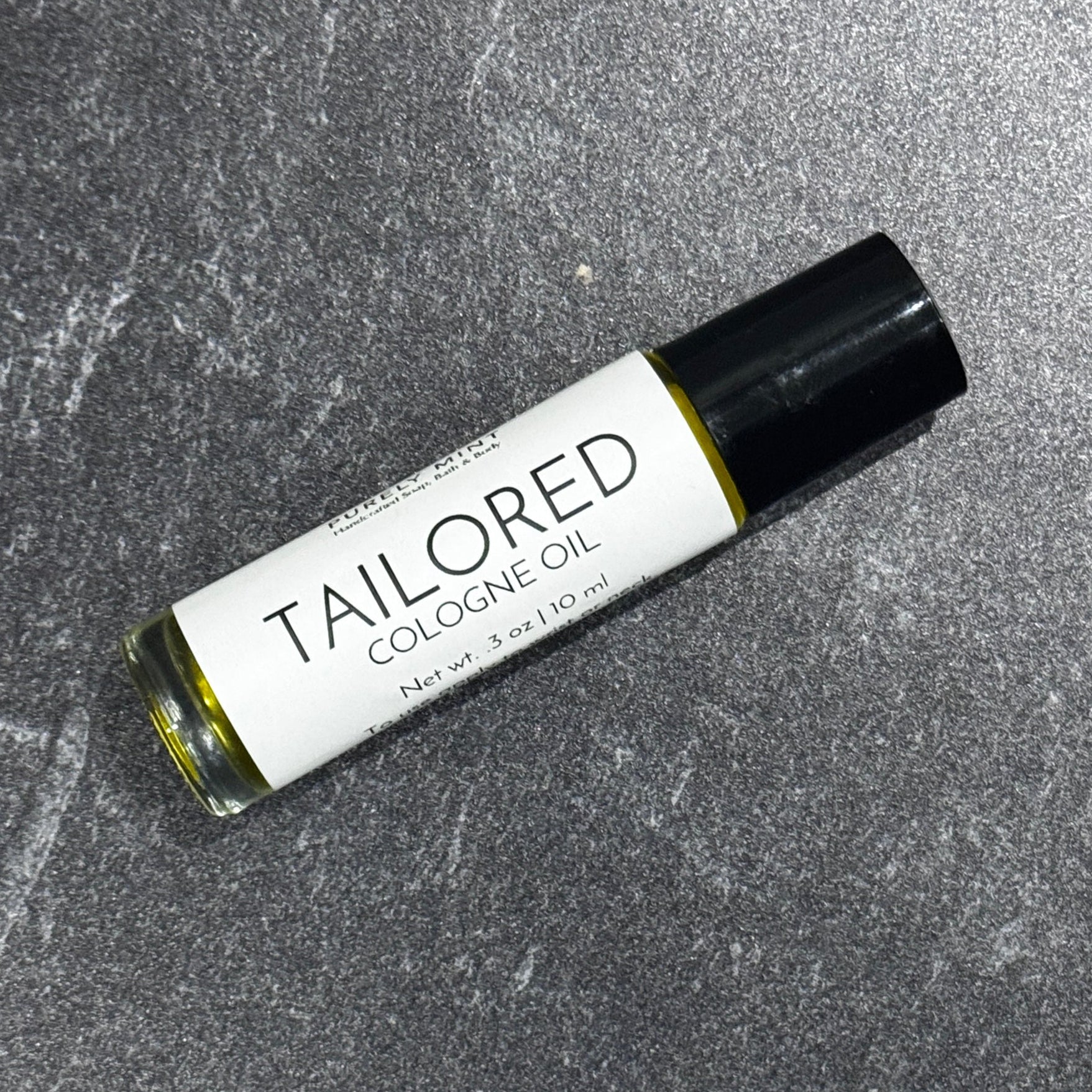 A 10 ml roll-on bottle of Tailored Cologne Oil by Purely Mint, featuring a white label and black cap, resting on a textured gray stone surface.