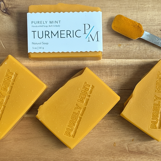 Three bars of yellow turmeric soap and one wrapped bar with a white label reading 'Turmeric Natural Soap' are arranged on a wooden surface, with a small metal scoop of turmeric powder beside them.