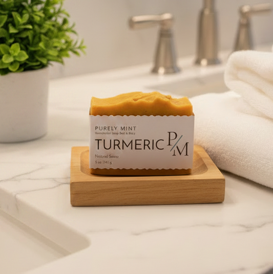 A bar of handmade Purely Mint Turmeric Soap sits on a small wooden soap dish on a white marble bathroom counter, next to a potted green plant and folded white towels.