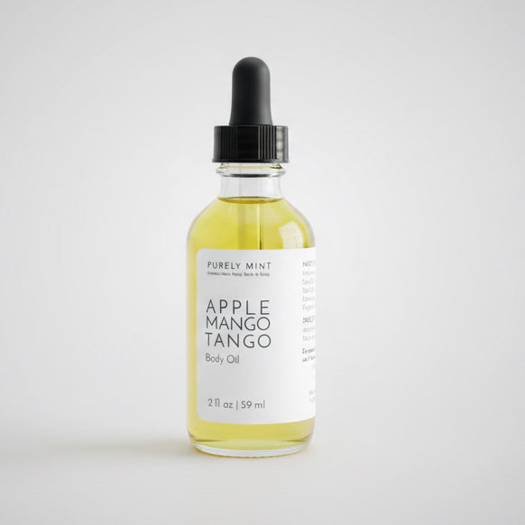 A bottle of Apple Mango Tango body oil is featured in a commercial photogray packshot. The background is a light gray seamless backdrop, and the lighting is neutral.