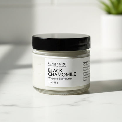 Black Chamomile whipped body butter jar in a modern bathroom, white marble counter, green plant.