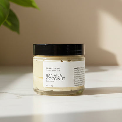 Jar of banana coconut body butter on a white marble counter with a green plant.