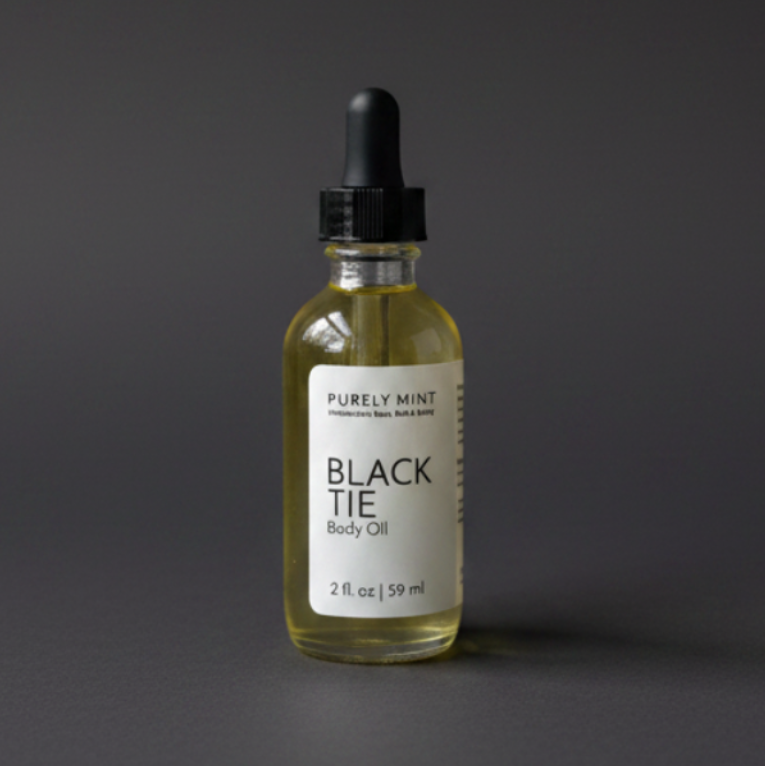 A bottle of Purely Mint Black Tie Body Oil with pale yellow oil inside, a white label, and a black dropper top, set against a dark gray backdrop.