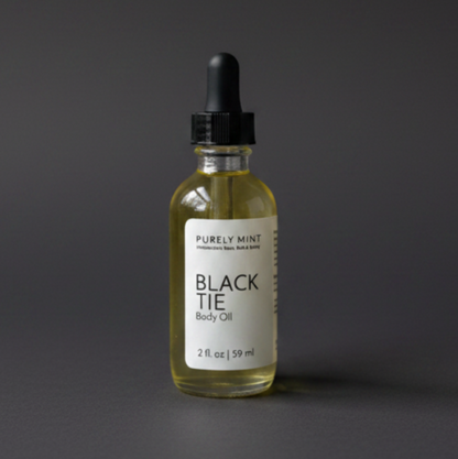 A bottle of Purely Mint Black Tie Body Oil with pale yellow oil inside, a white label, and a black dropper top, set against a dark gray backdrop.