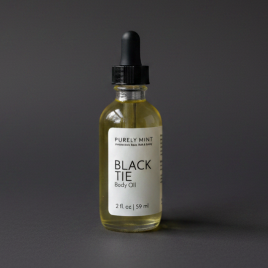 A bottle of Purely Mint Black Tie Body Oil with pale yellow oil inside, a white label, and a black dropper top, set against a dark gray backdrop.