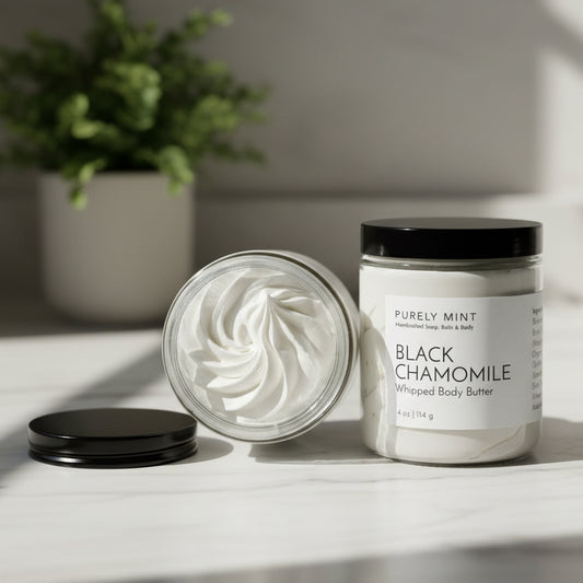 Black Chamomile whipped body butter jar with lid in a modern bathroom, white marble counters, green plant .