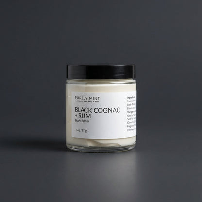 2 oz Black cognac & rum body butter in a clear glass jar with a white label and sleek black top on a dark gray surface. 