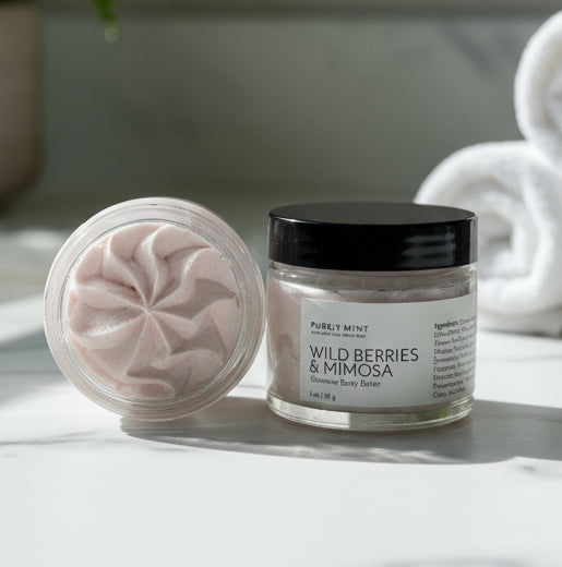 Two small jars of wild berries & mimosa  body butter with labels on a white marble counter with rolled washcloths and a green plant.