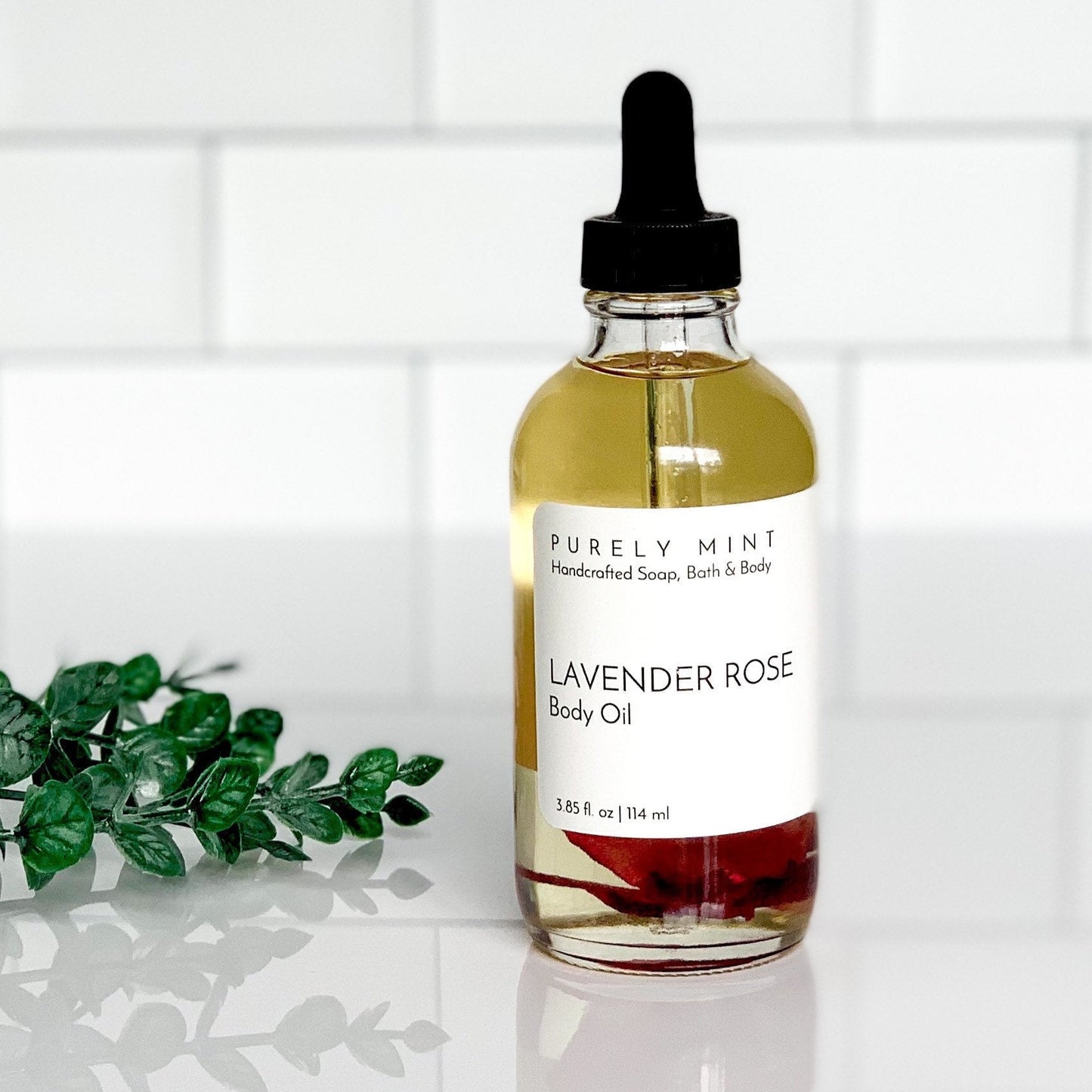 A 4 oz Bottle of Lavender Rose Natural body oil with a white label and dropper top on a white surface with green leaves and a white tiled background.