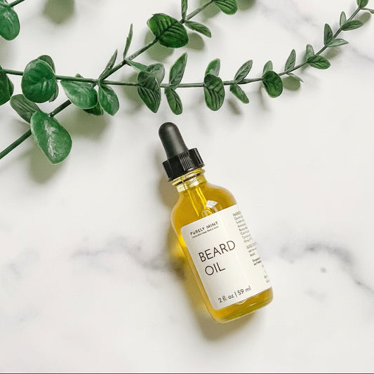 A 2 oz glass bottle of Gentlemen's Beard Oil with a dropper, placed on a marble surface with a green plant in the background.